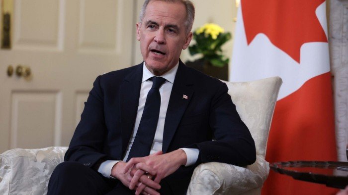 Mark Carney in a suit and sits in an armchair in front of a Canadian flag