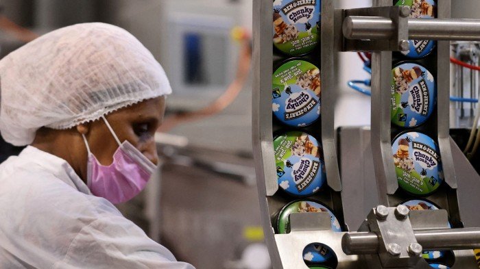 A worker in a hairnet and pink mask operates a production line at a Ben & Jerry's factory, with ice cream pots labelled "Chunky Monkey" visible on the conveyor