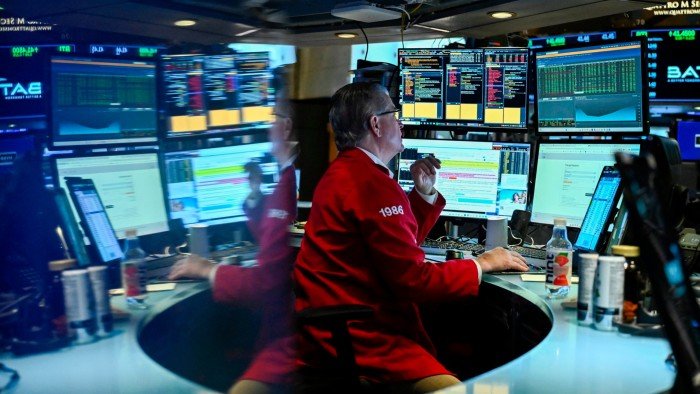 A trader in a red jacket operates multiple computer screens on the New York Stock Exchange floor,