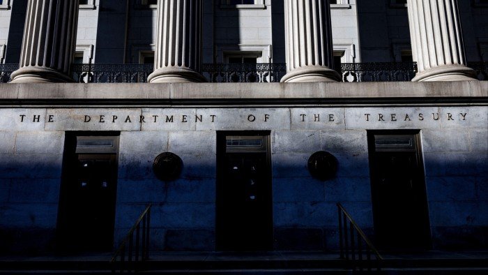 The front facade of the US Treasury building in Washington with tall columns and the inscription "THE DEPARTMENT OF THE TREASURY." Shadows partially cover the lower portion of the building.