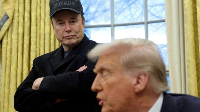 Elon Musk, standing, looks at Donald Trump, seated, who is speaking, in the Oval Office