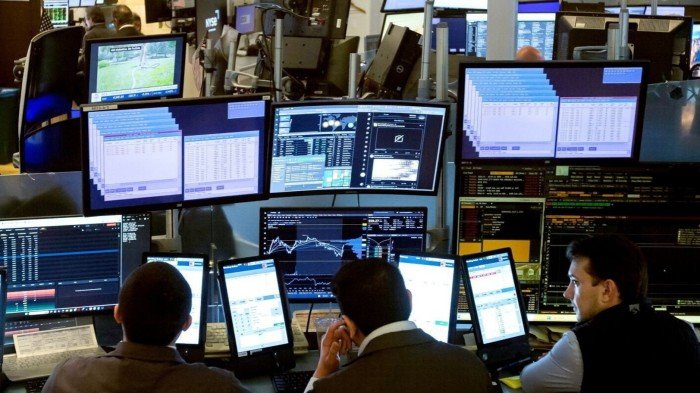 Traders are seated at desks on the New York Stock Exchange floor, surrounded by multiple computer screens displaying financial data and stock charts